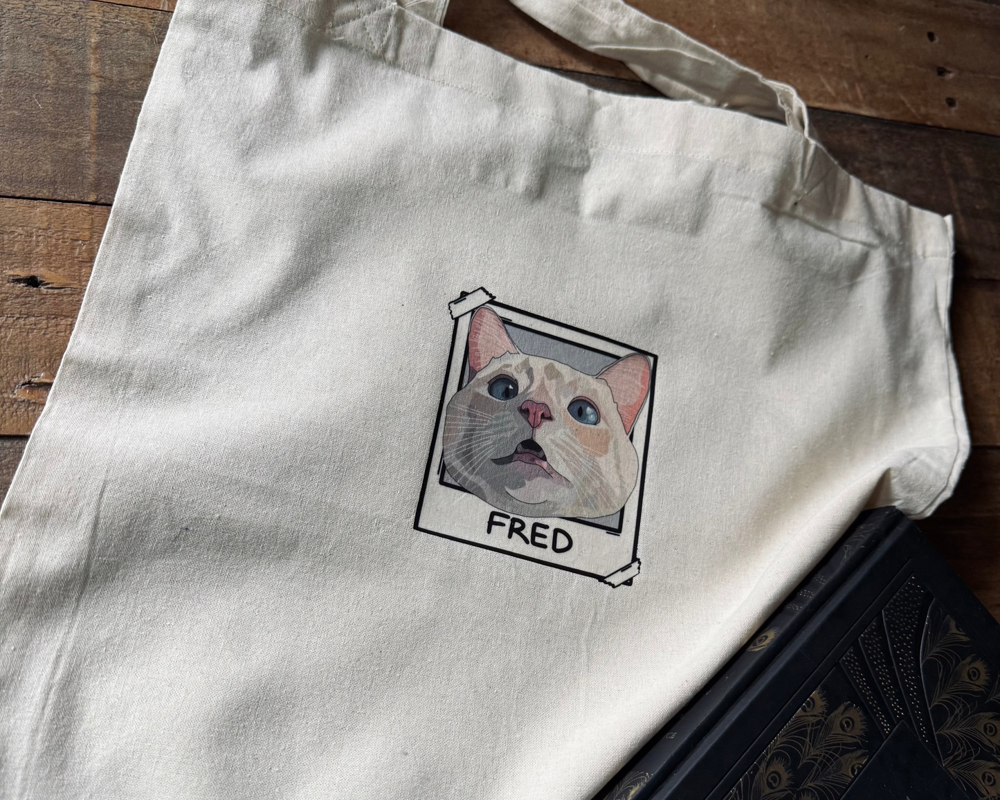 White tote bag with a cat illustration and 'FRED' text on a wooden surface