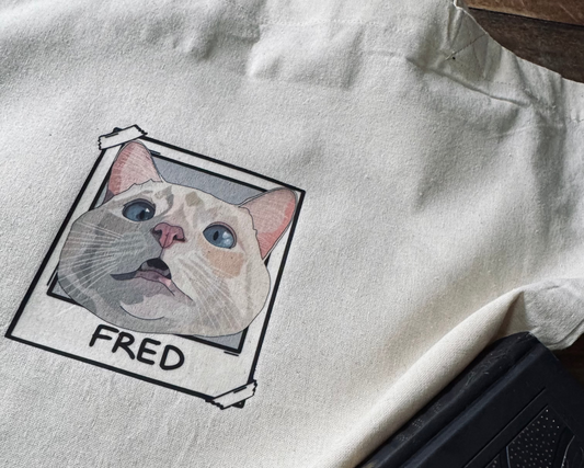 White tote bag with a cat illustration and 'FRED' text on a wooden surface