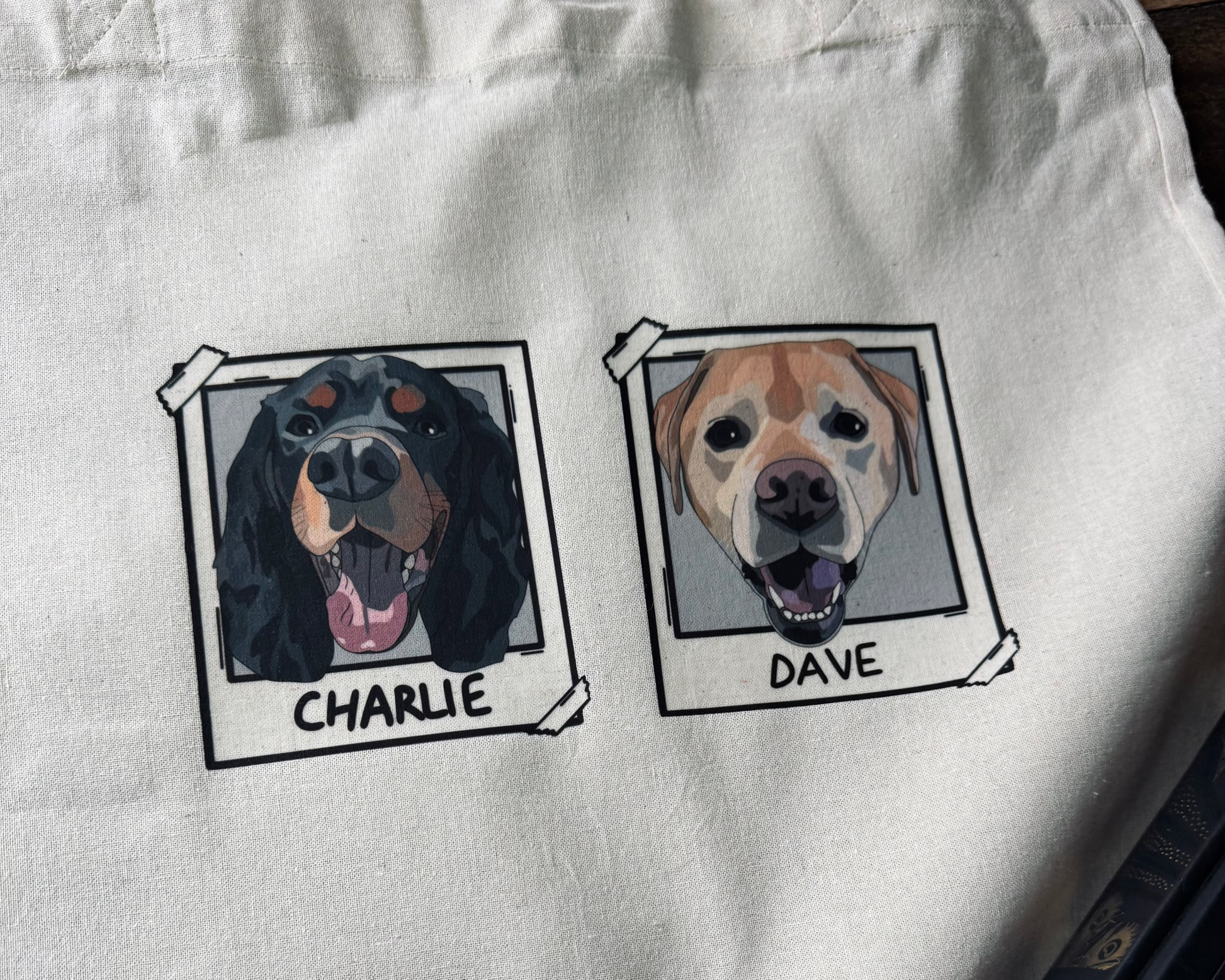 White tote bag with illustrations of two dogs named Charlie and Dave on a wooden surface.