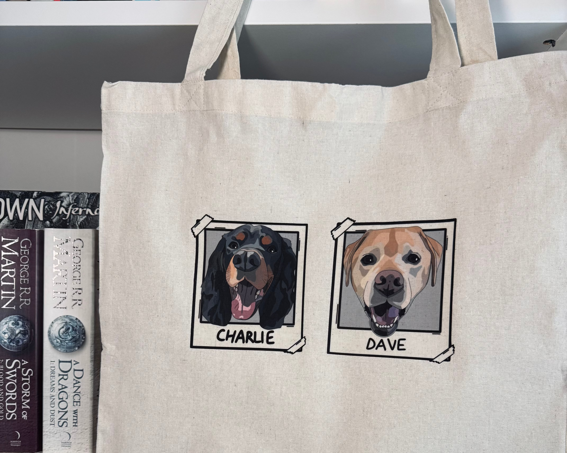 Beige tote bag with illustrations of two dogs named Charlie and Dave, placed on a surface with books in the background.