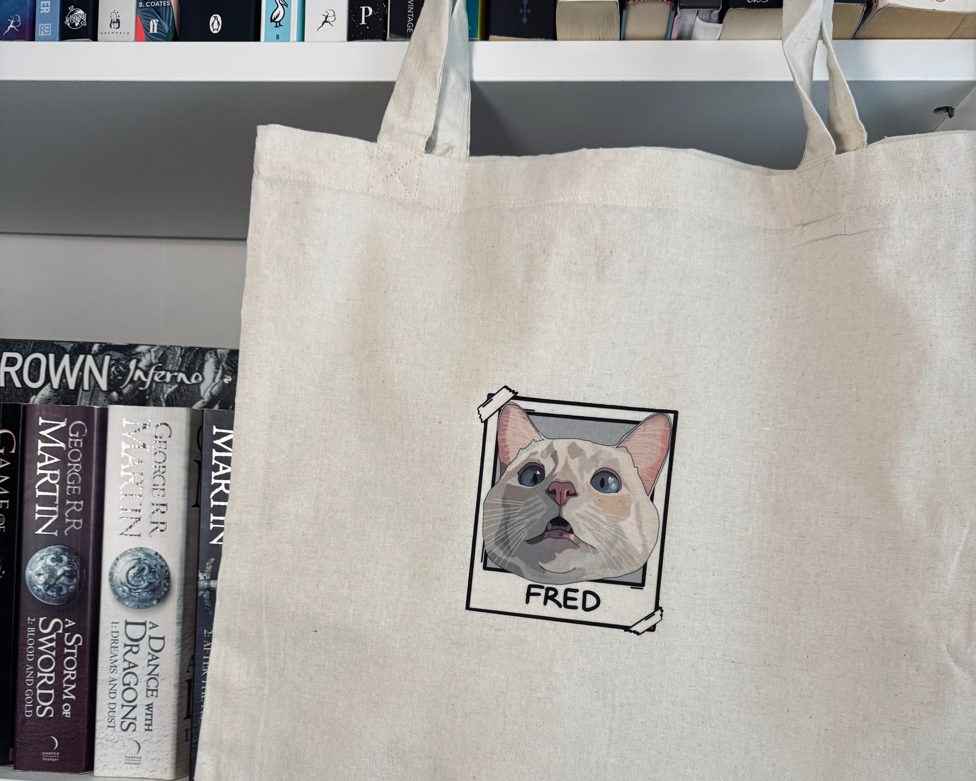 Beige tote bag with a cat illustration and 'FRED' text, placed on a shelf with books.