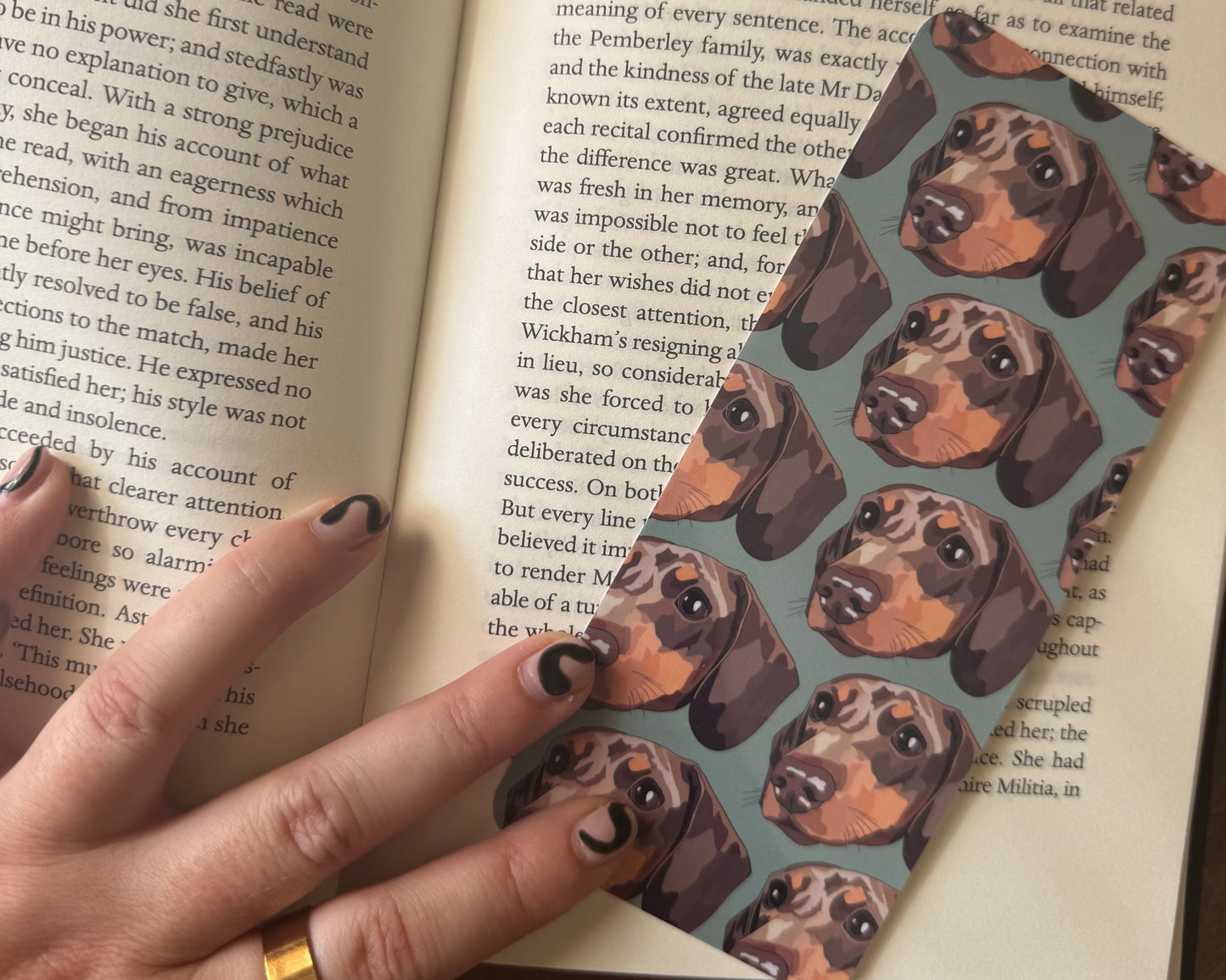 Open book with a hand holding a bookmark featuring dachshund dog pattern.